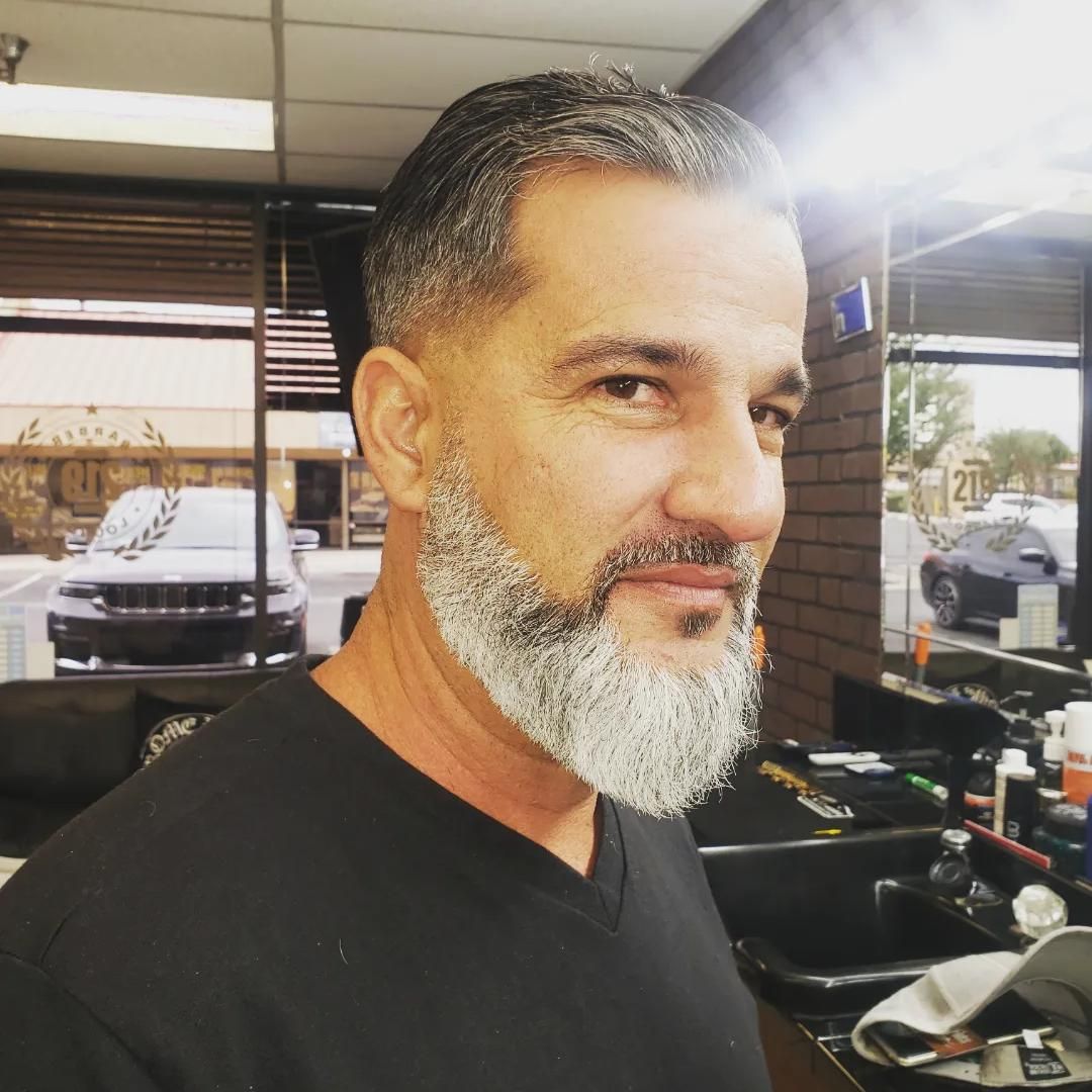 Man with gray beard in a barbershop, looking sideways with a slight smile.