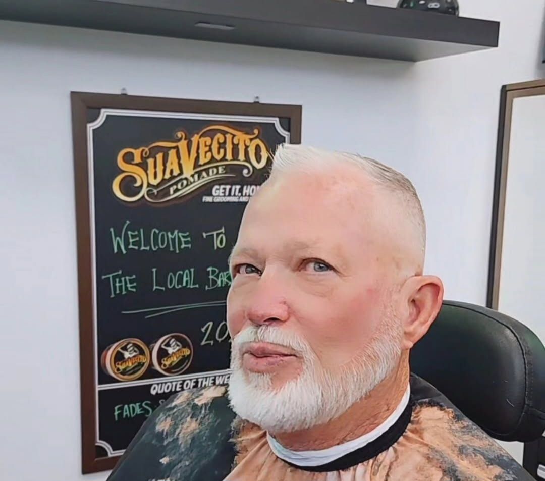 Smiling bald man with white beard in a barber chair, with a chalkboard sign behind him.