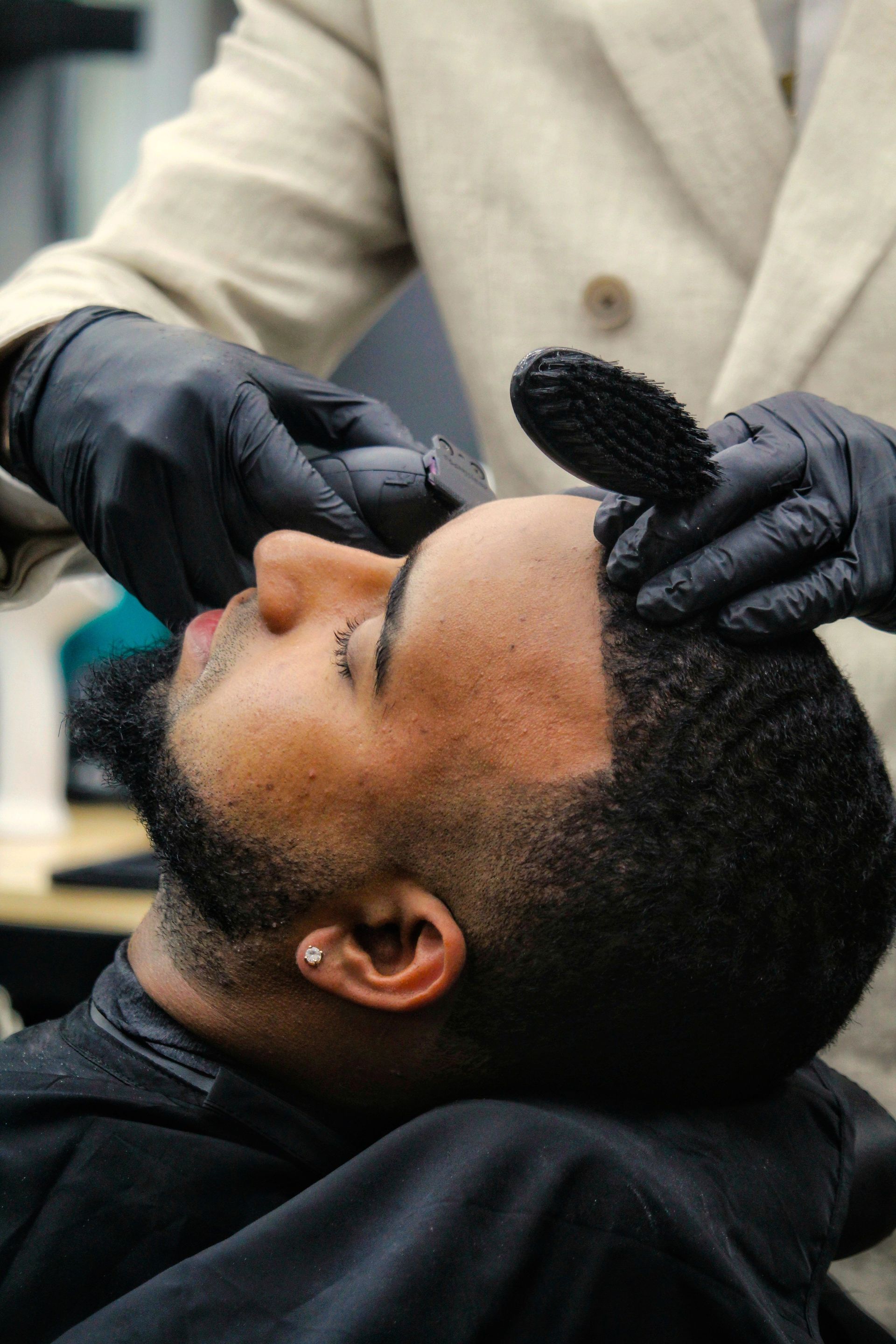 Gloved barber trimming a man's beard with a brush and clipper in a salon chair