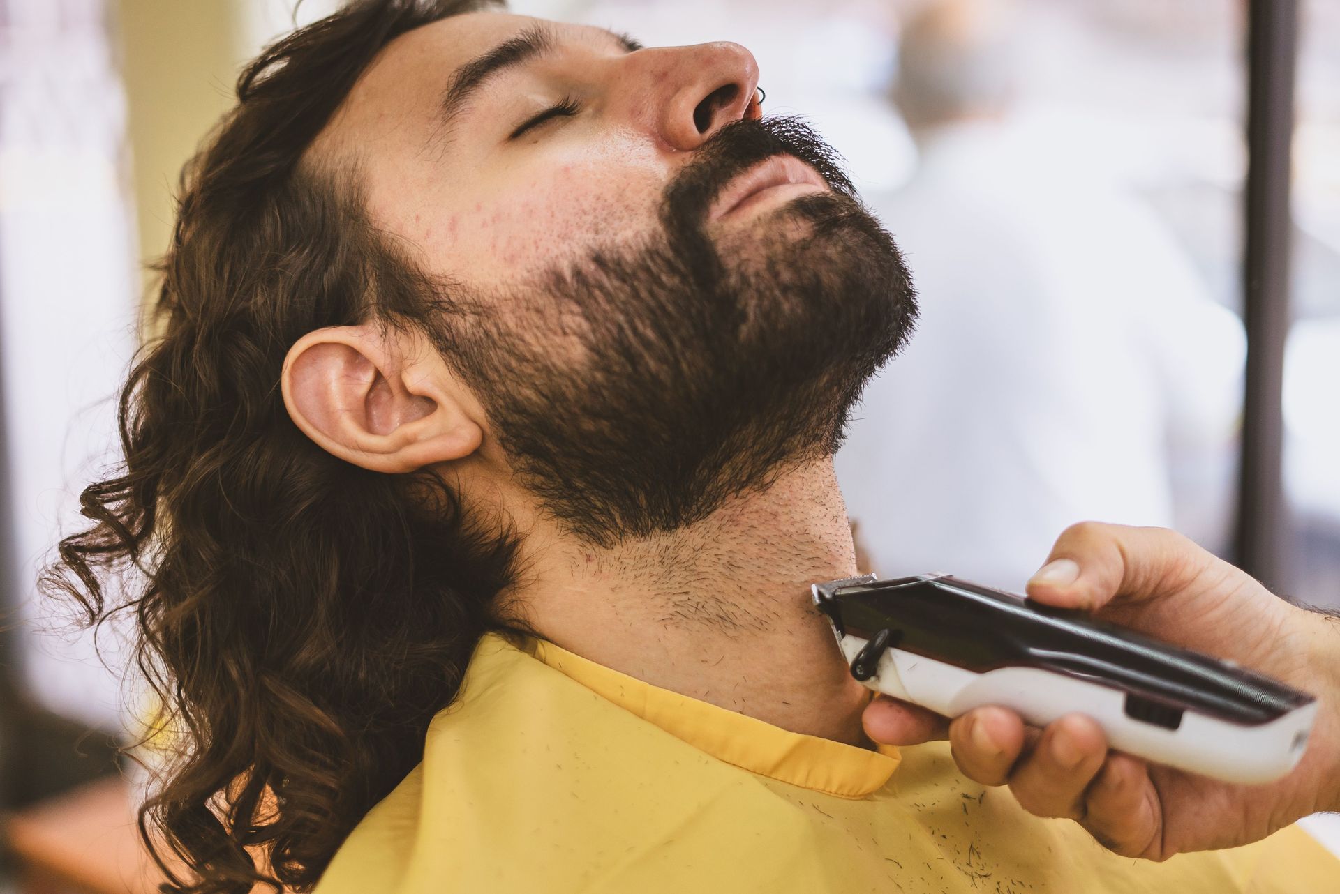 Bearded man with eyes closed getting his neck shaved by a barber with electric clippers