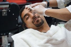 Man reclining in a barber chair while a stylist applies shaving cream to his face