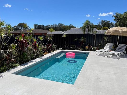 Swimming Pool With a Waterfall Feature, Patio, and Glass Fencing — Clearwater Pools Hallidays Point in Hallidays Point, NSW