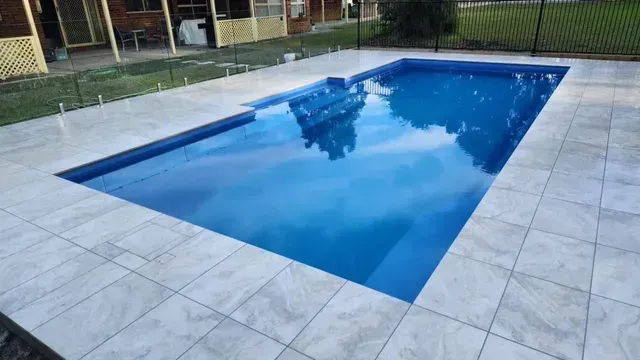 Backyard swimming pool with blue water, surrounded by light tile patio and a house in the background