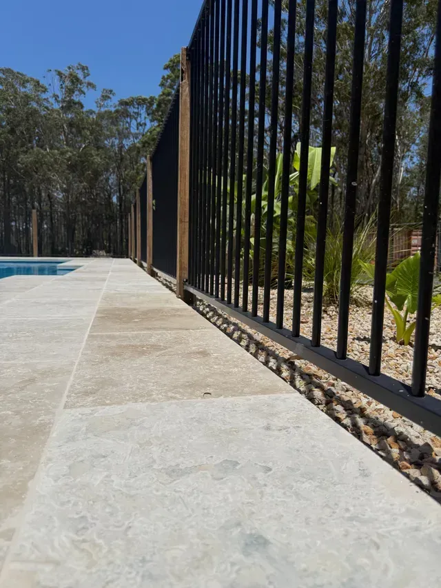 Concrete poolside walkway beside a black metal fence and trees in bright sunlight