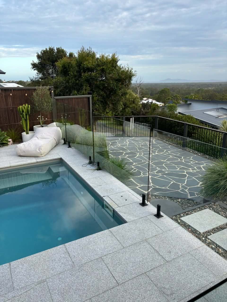 Poolside patio with glass railing, white loungers, and ocean view beyond a landscaped yard — Clearwater Pools Hallidays Point in Hallidays Point, NSW