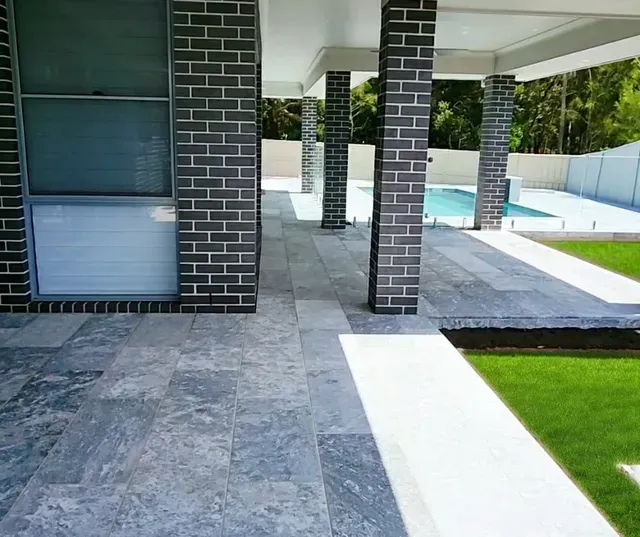 Covered outdoor patio with gray stone flooring, brick columns, and a strip of green lawn
