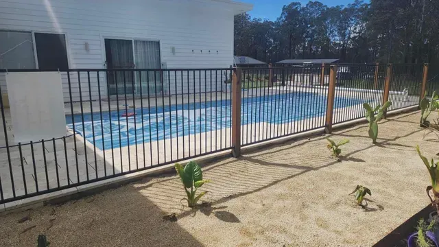 Backyard pool behind black metal fence beside a white house under a clear sky