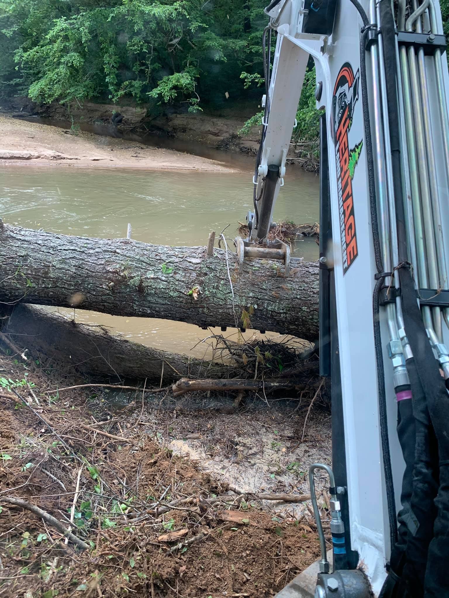 Excavator removing a log from a stream. The machine is white with orange accents, and the water is murky.