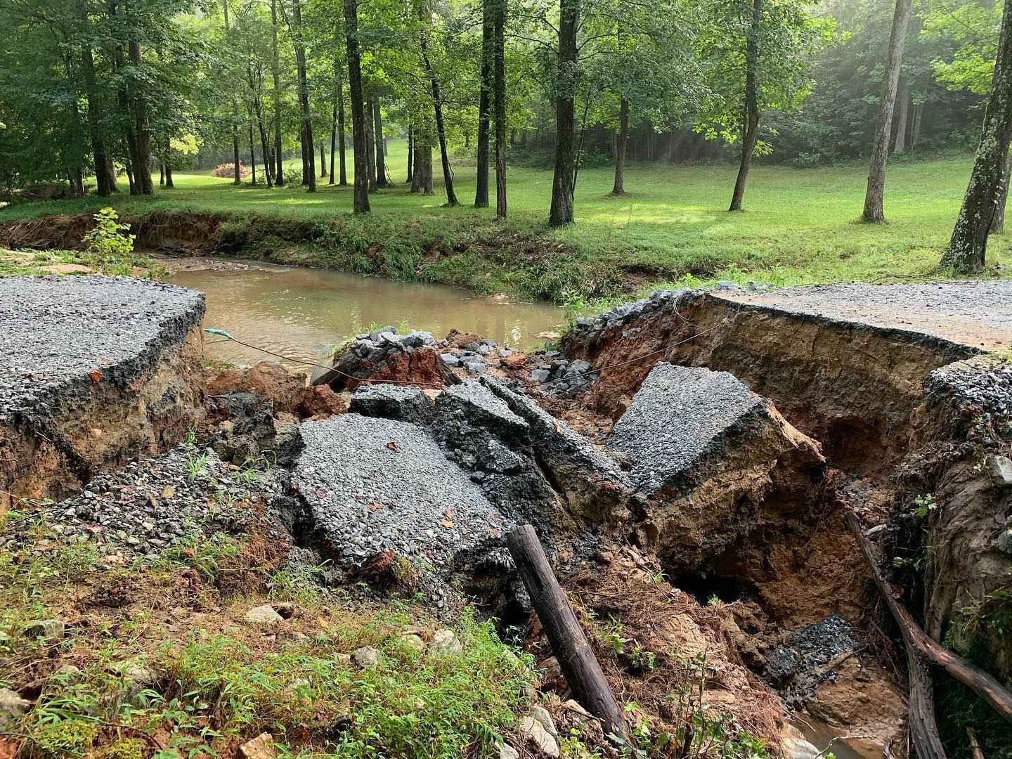Road collapsed, showing exposed earth and gravel, spanning a creek with trees in background.