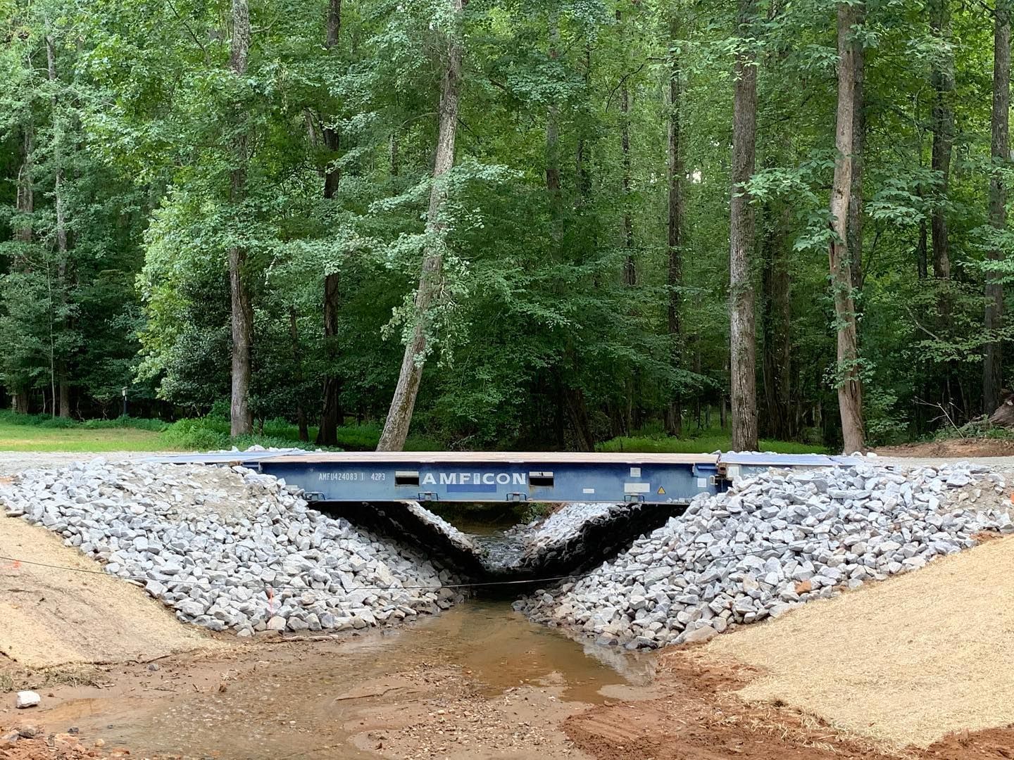 A small bridge over a creek in a forest. Gray steel bridge, stone embankments, trees in background.
