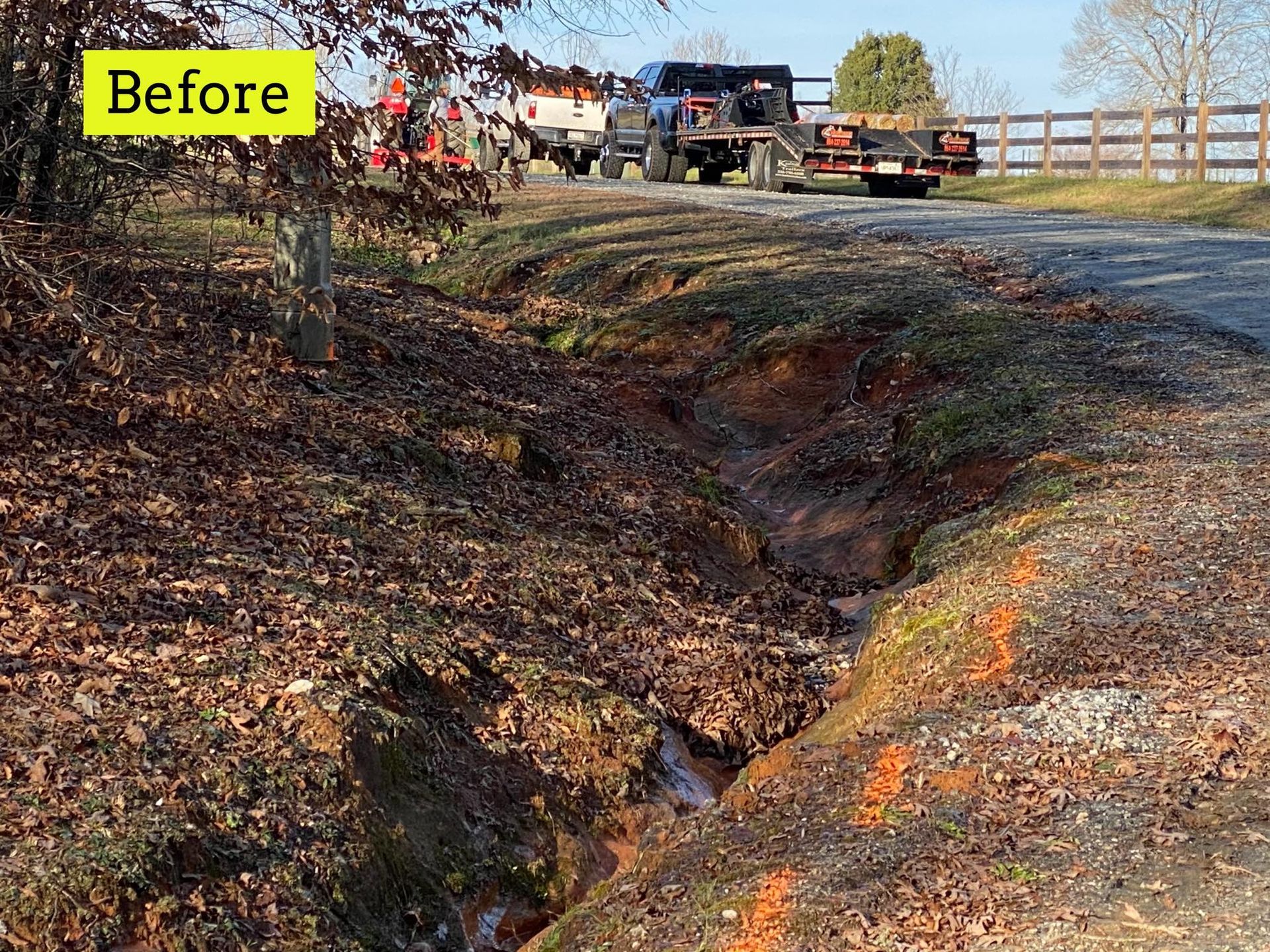 Eroded ditch next to a road, before construction. Brown earth and foliage. Trucks and fence in the background.