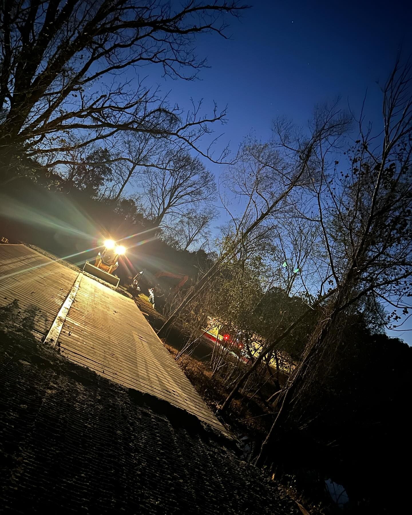 Construction vehicle with bright lights on a roof at dusk, silhouetting trees against a blue sky.