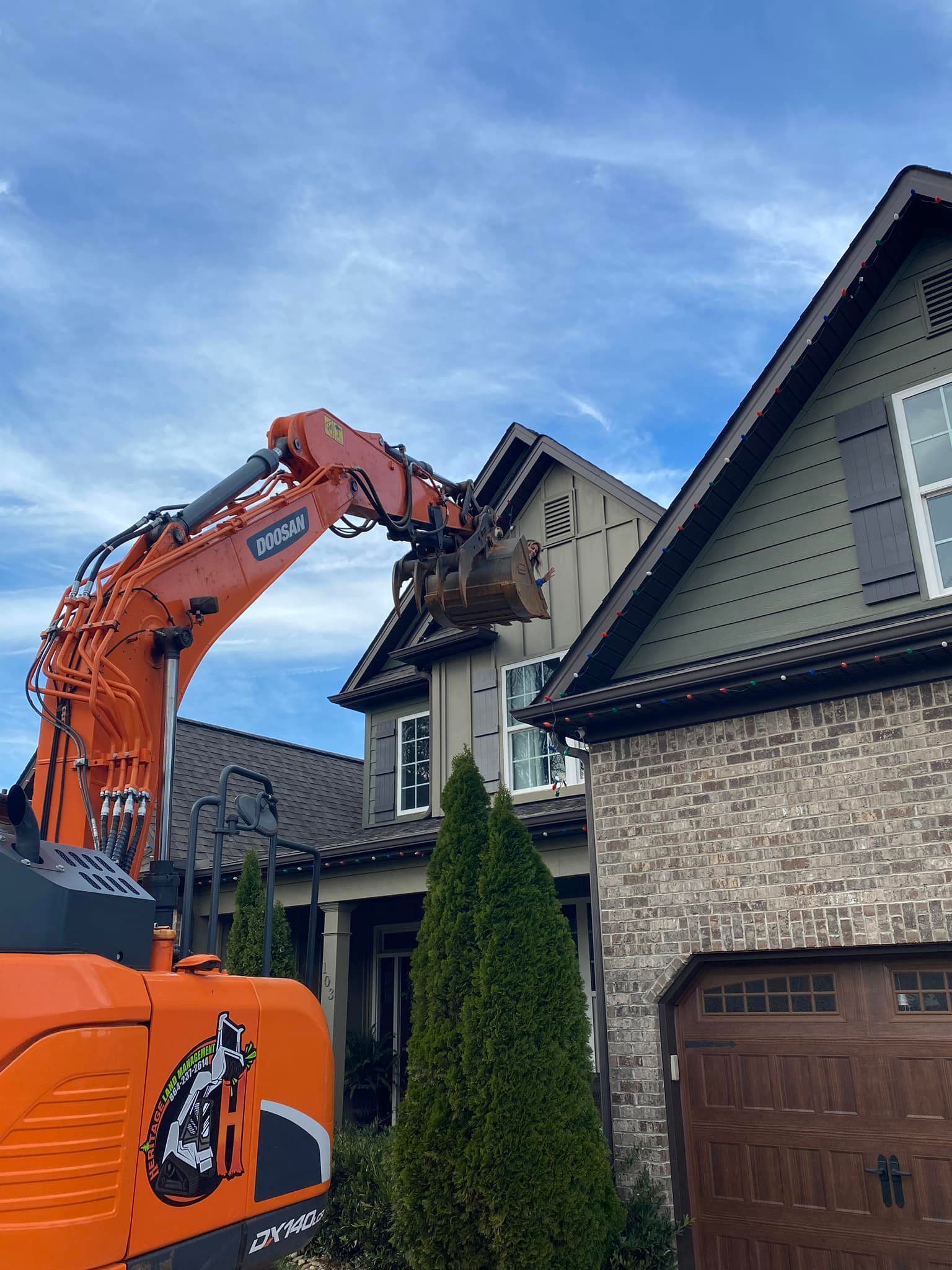 An orange excavator demolishes a two-story house, while another house stands next to it. Blue sky.