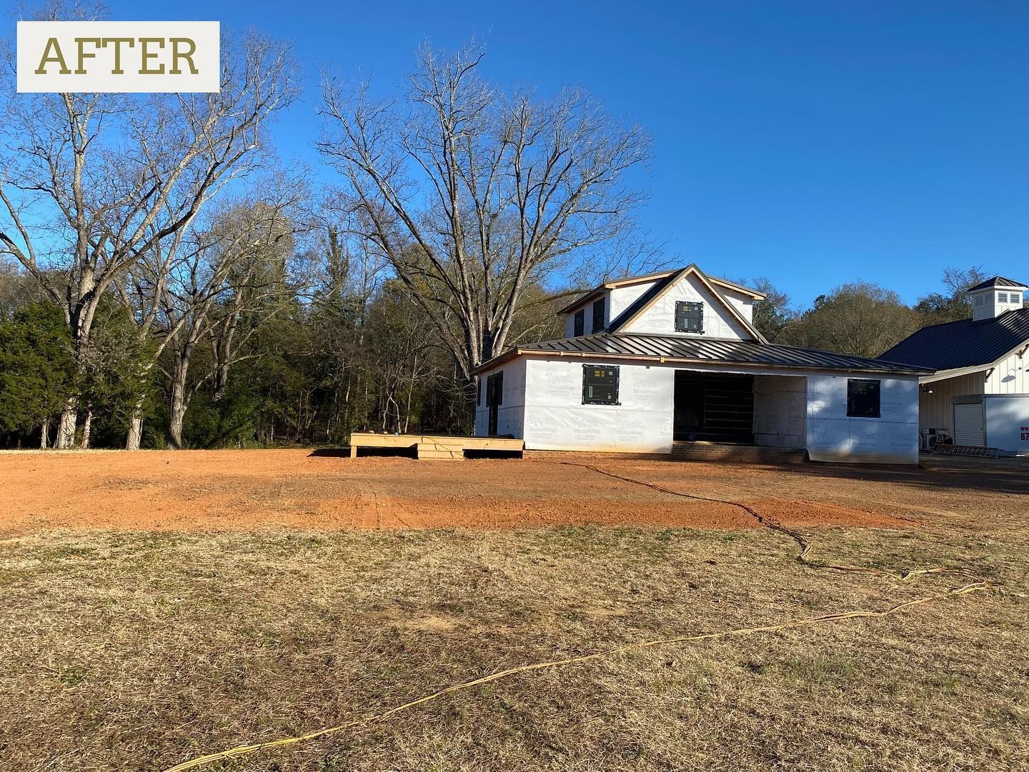 White farmhouse with black trim, under construction, set in a field. Trees and blue sky in background.