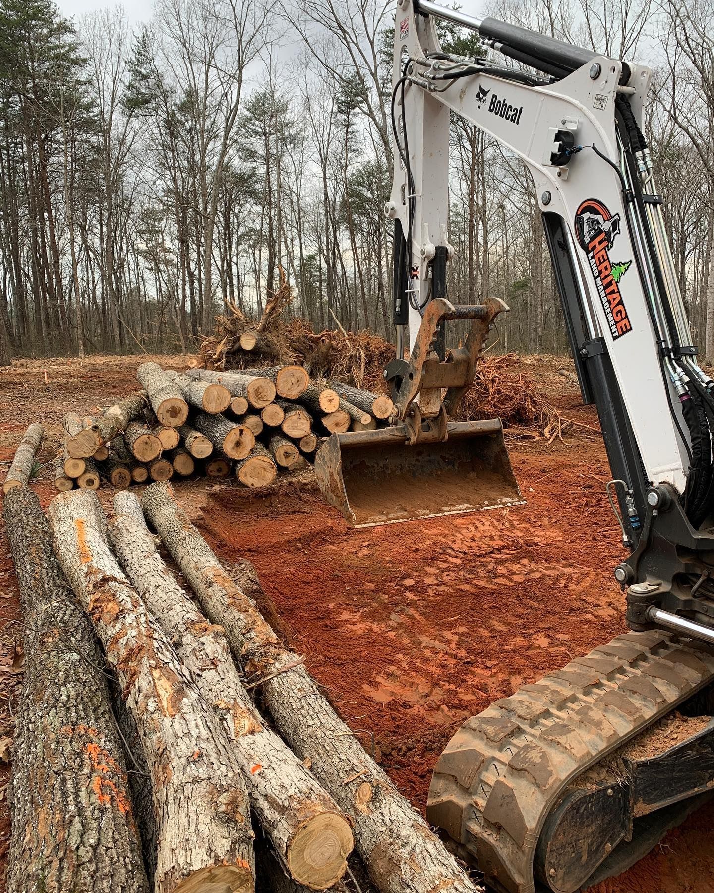 Excavator moving logs on a red dirt clearing. White machine, brown logs, trees in background.