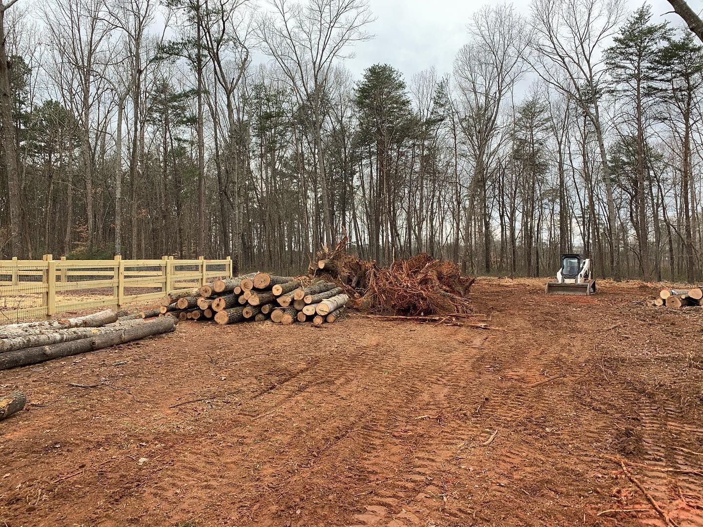 Cleared area with logs, brush pile, and small loader; trees and fence in background.