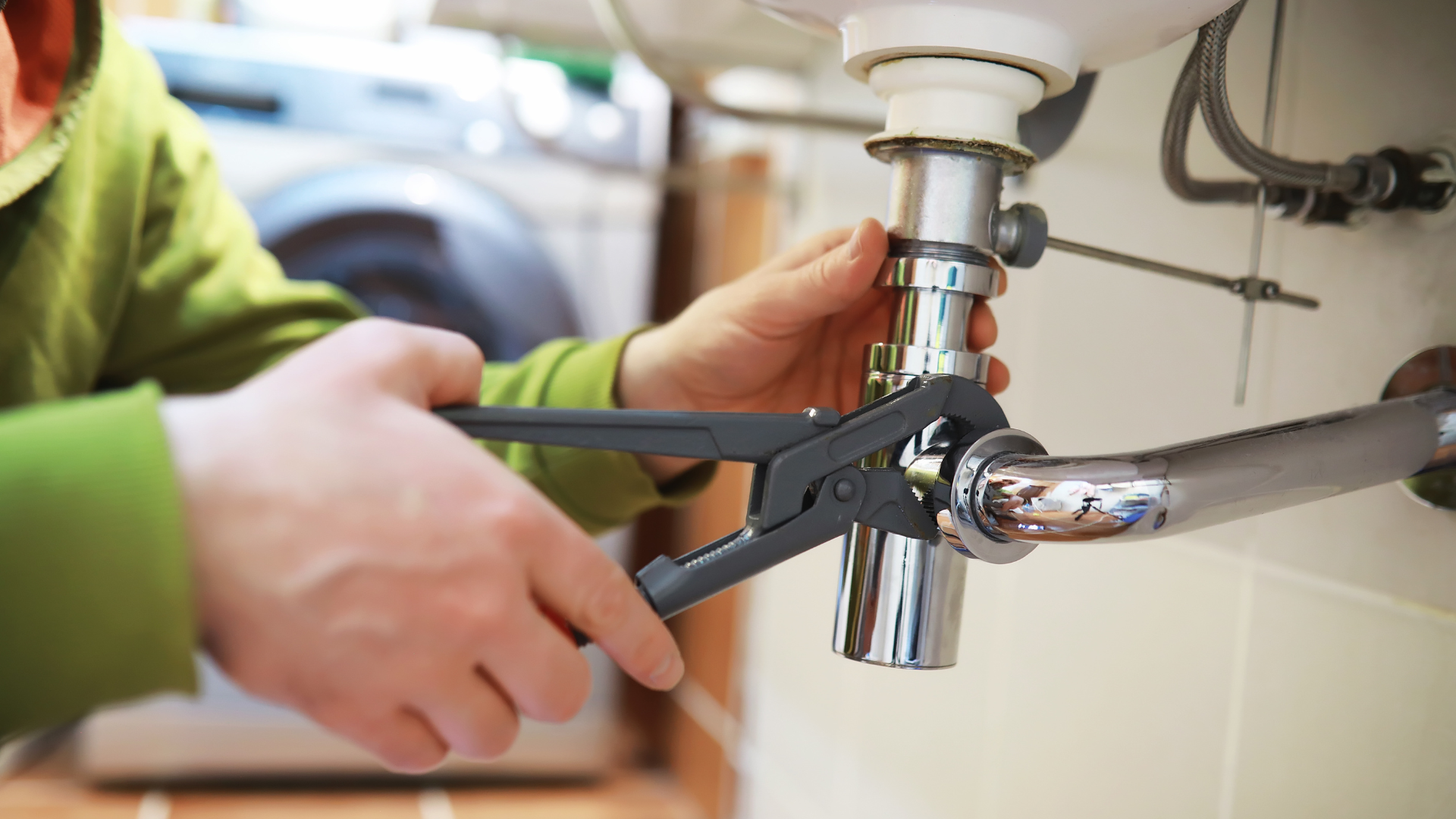 Plumber using pliers to tighten pipes under a white sink in a bathroom.