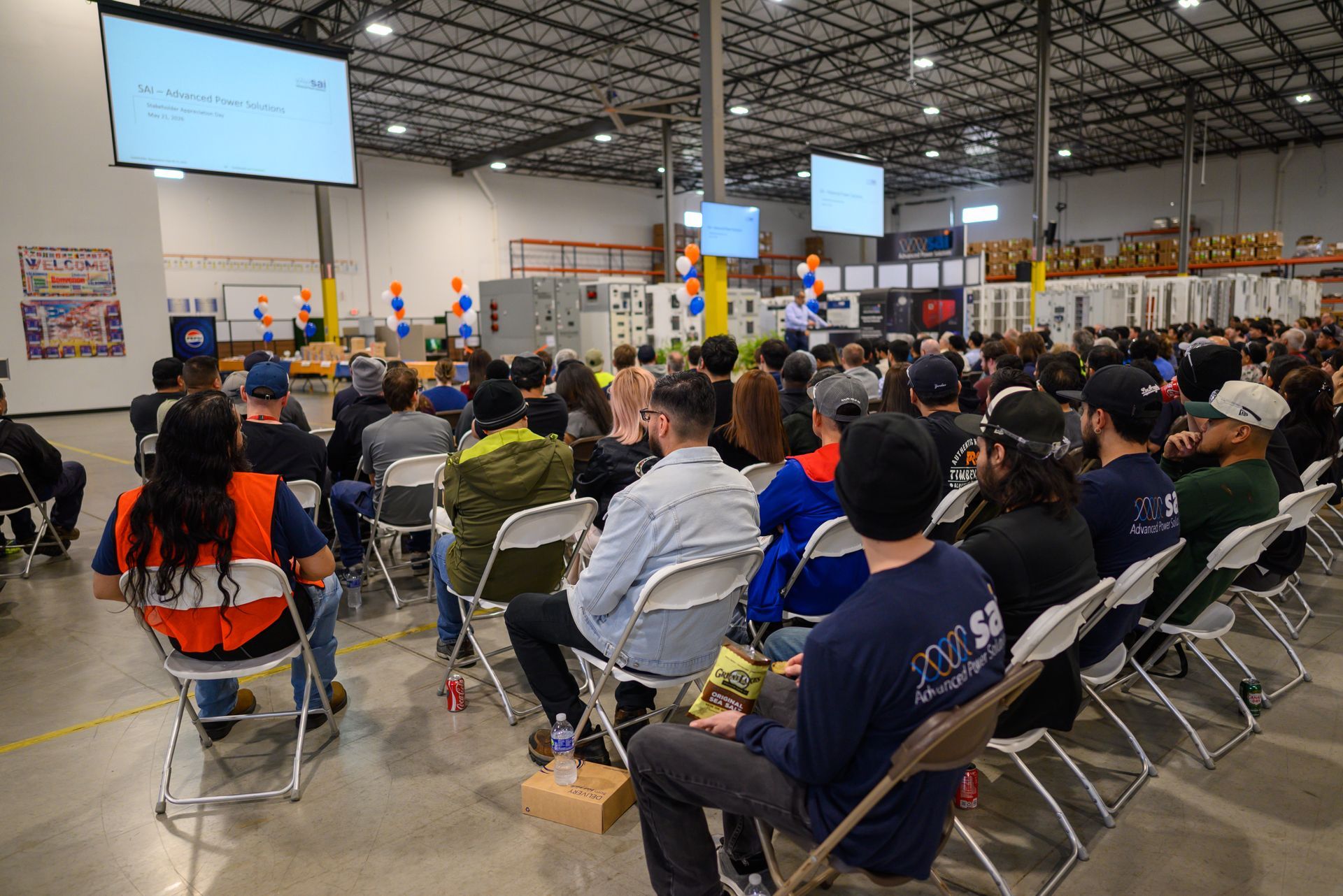 People seated in a large warehouse listening to a presentation on screens at the front