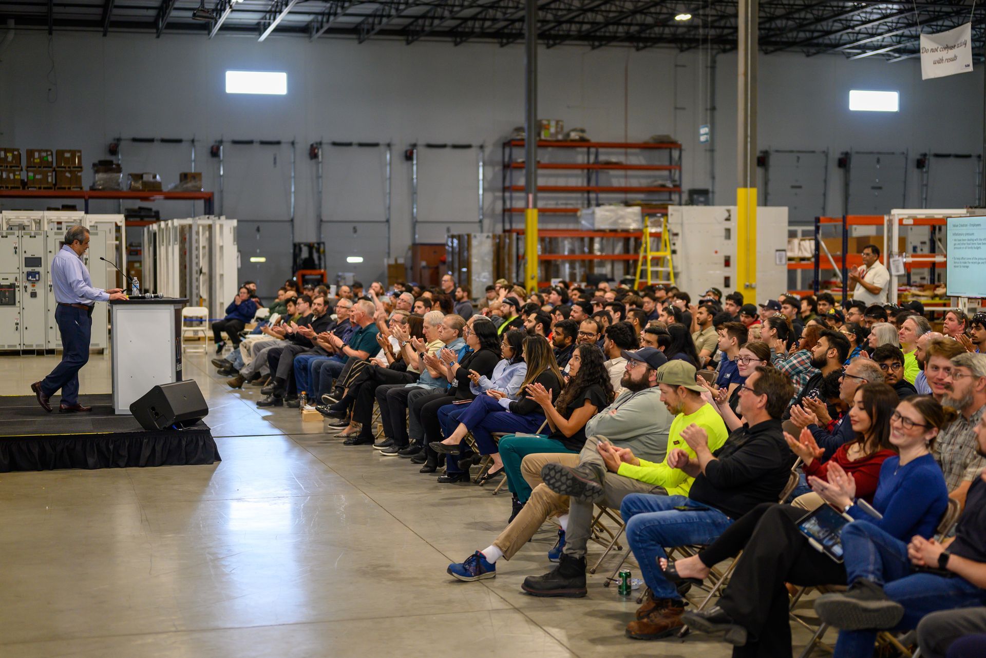 Audience seated in warehouse listening to a speaker at a podium beside a stage