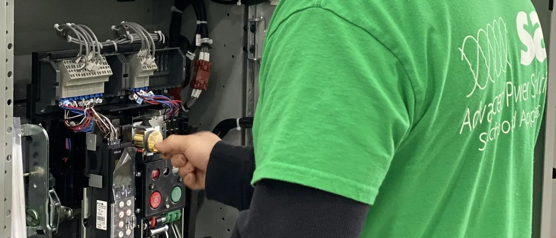 Person in a green shirt working on an open electrical panel with tools inside a utility room