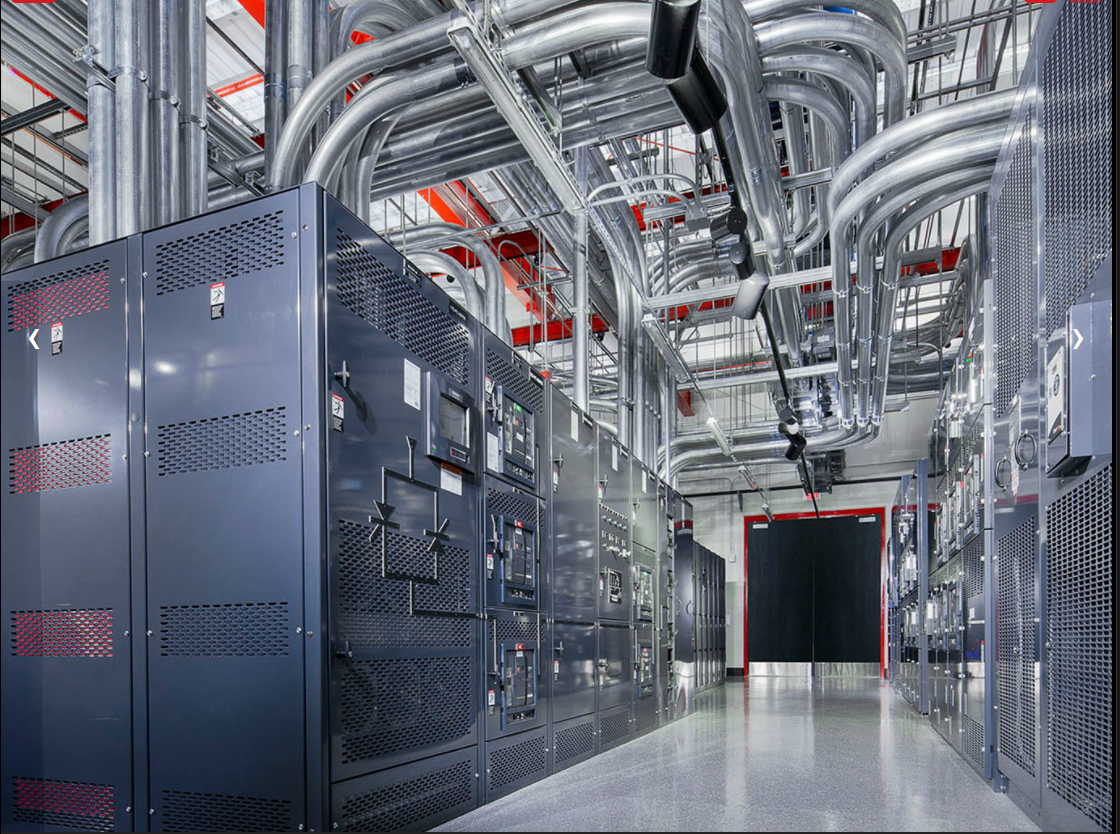 Server room with rows of black cabinets and silver overhead cables in a bright industrial data center
