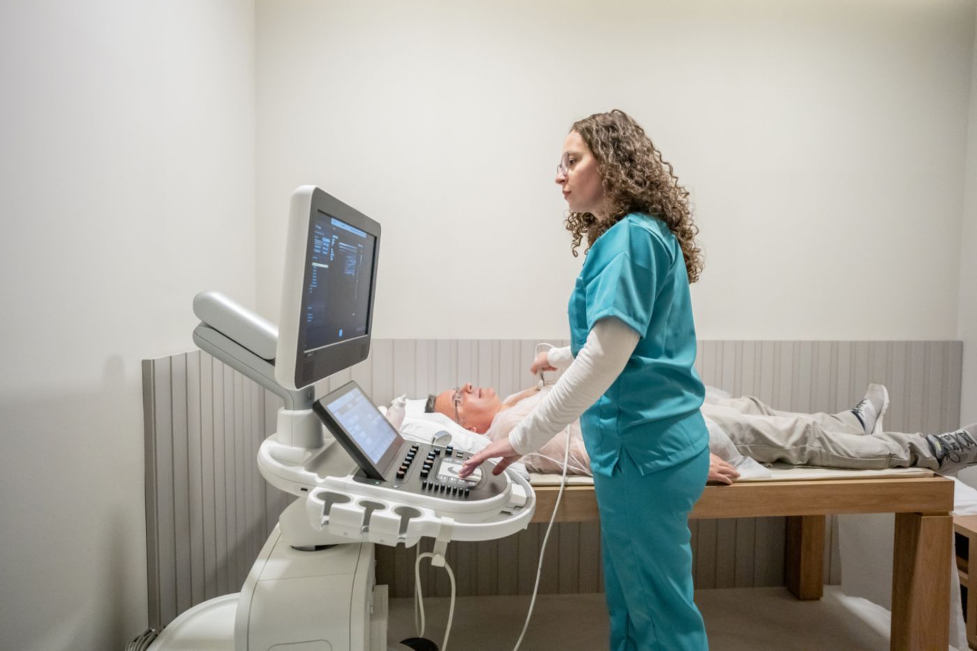Sonographer in teal scrubs performing an ultrasound on a patient in a clinic exam room