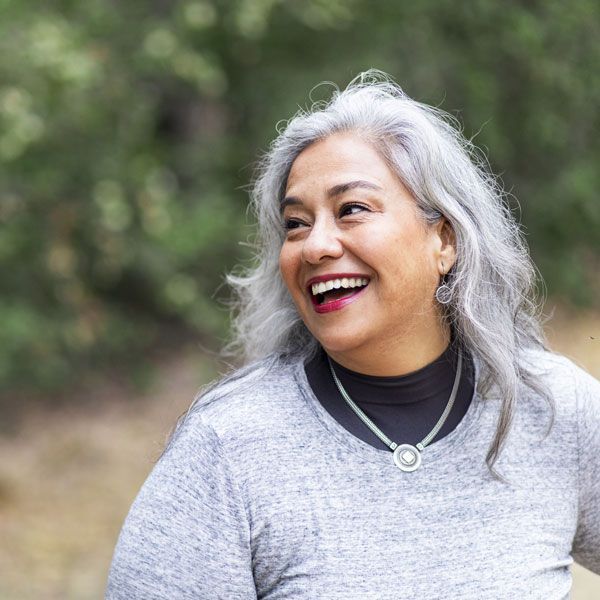 Smiling woman with gray hair in a gray top, standing outdoors against a blurred green background