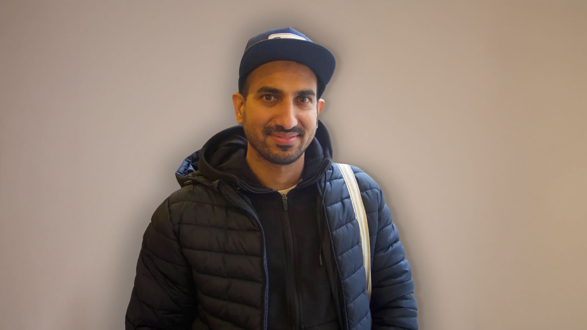 Mann i mørk jakke og baseballcaps smiler, skulderveske, nøytral bakgrunn.