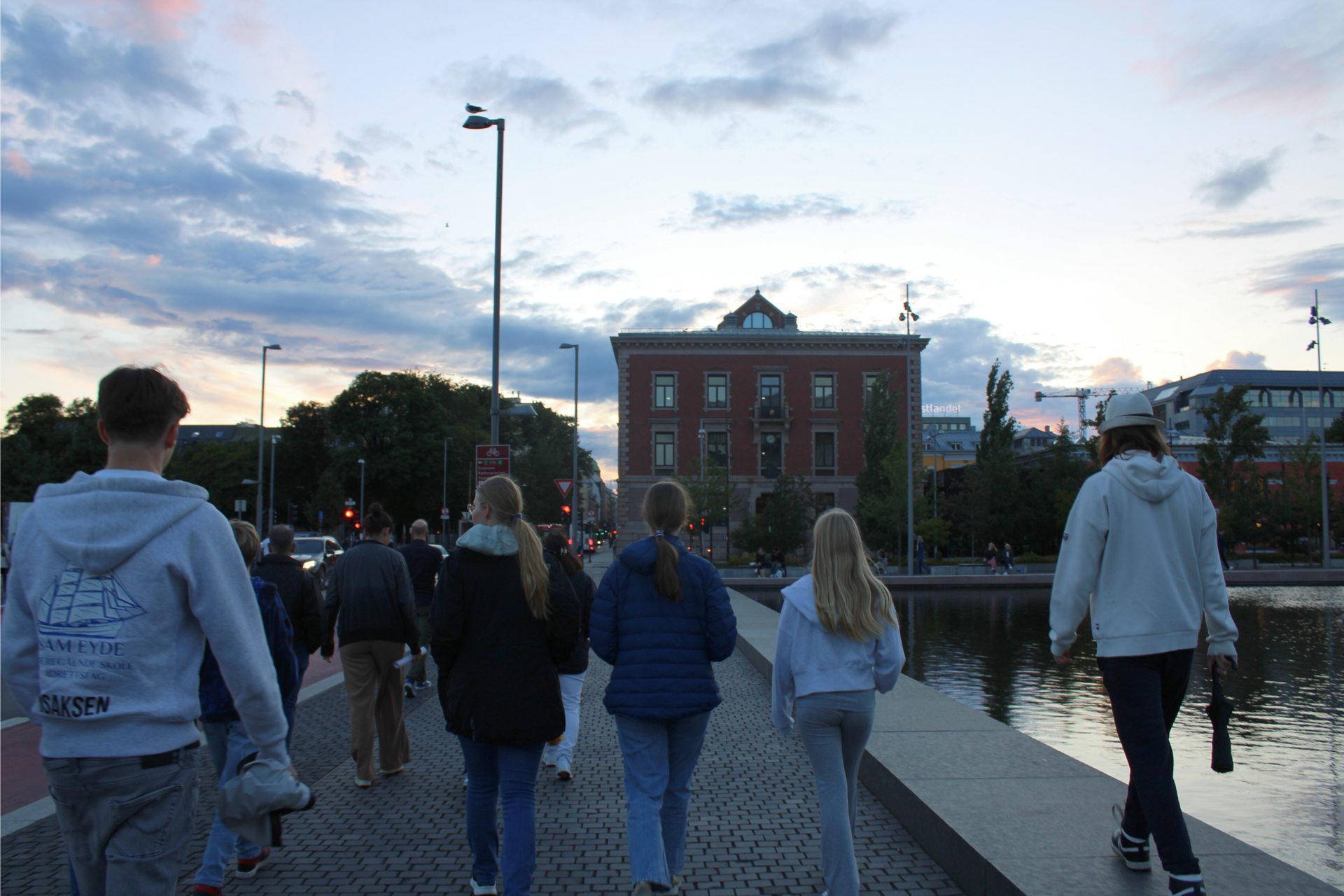 Folk går over en bro i skumringen. Rød mursteinsbygning i bakgrunnen, vann på siden.