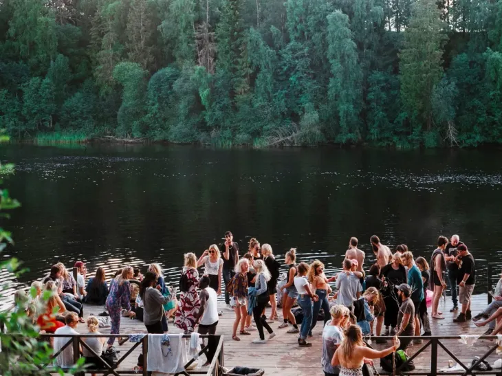 Folk samles på en trebrygge ved en innsjø med skog i bakgrunnen; solskinnsdag.