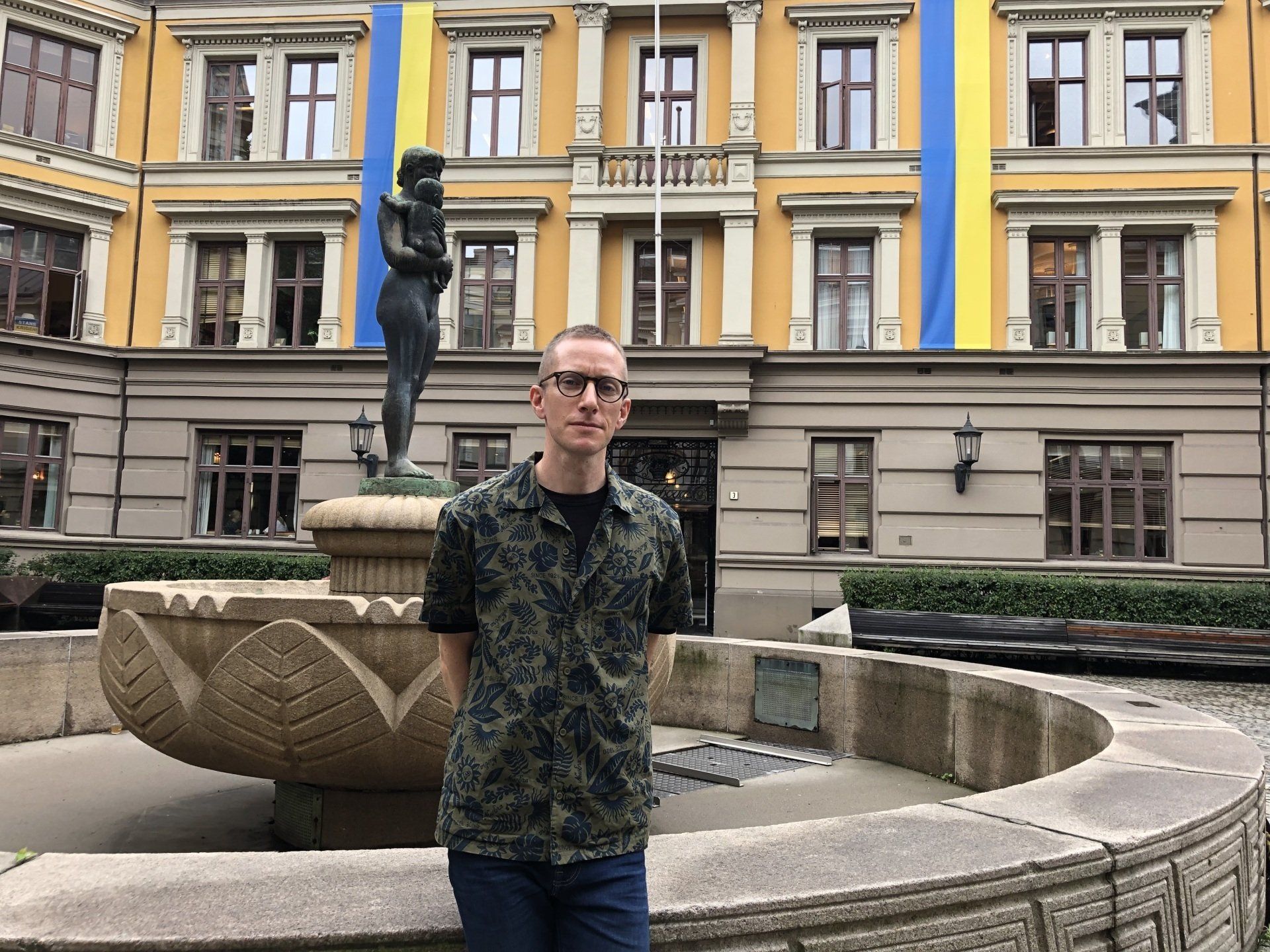 Mann i mønstret skjorte står ved en fontene foran en gul bygning med ukrainske flagg.