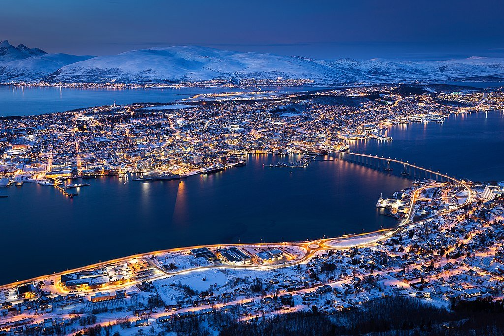 Bylys gløder om natten, snødekte bygninger ved en fjord, opplyst bro.