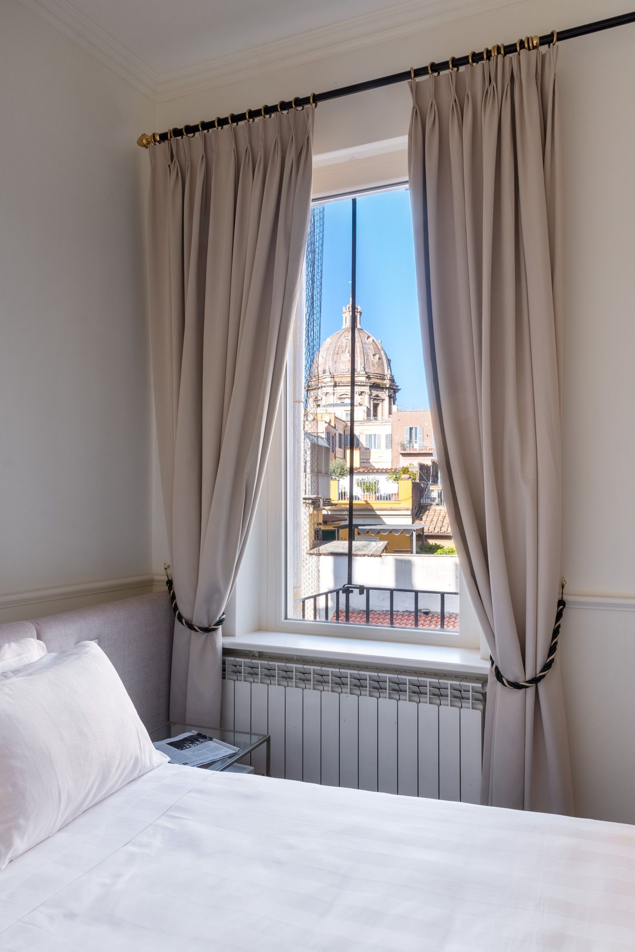 Bedroom window with view of a domed building, sunlight streams in. Beige curtains are tied back.