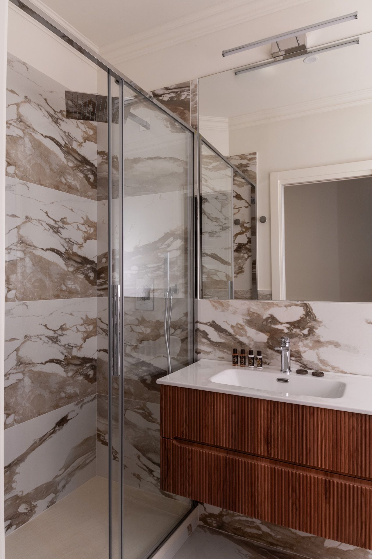 Bathroom with marble-look tile, glass shower, wood vanity, and a large mirror.