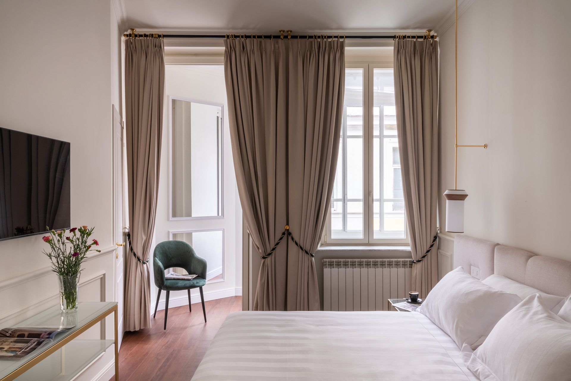 Bedroom with bed, window, and balcony. Beige curtains, white walls, wood floor, and a green chair.