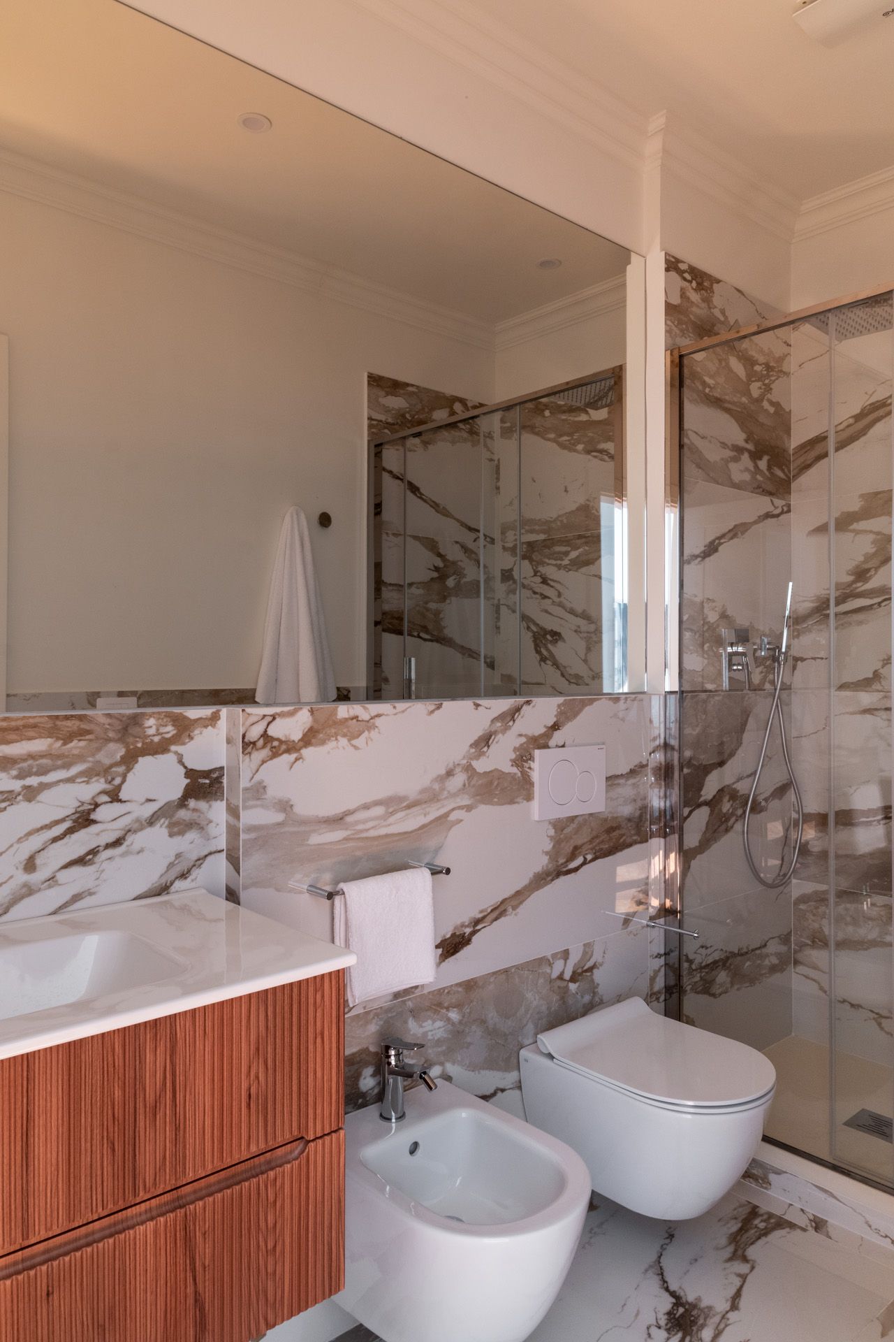 Luxury bathroom with marble walls, wooden vanity, large mirror, shower, toilet, and bidet.