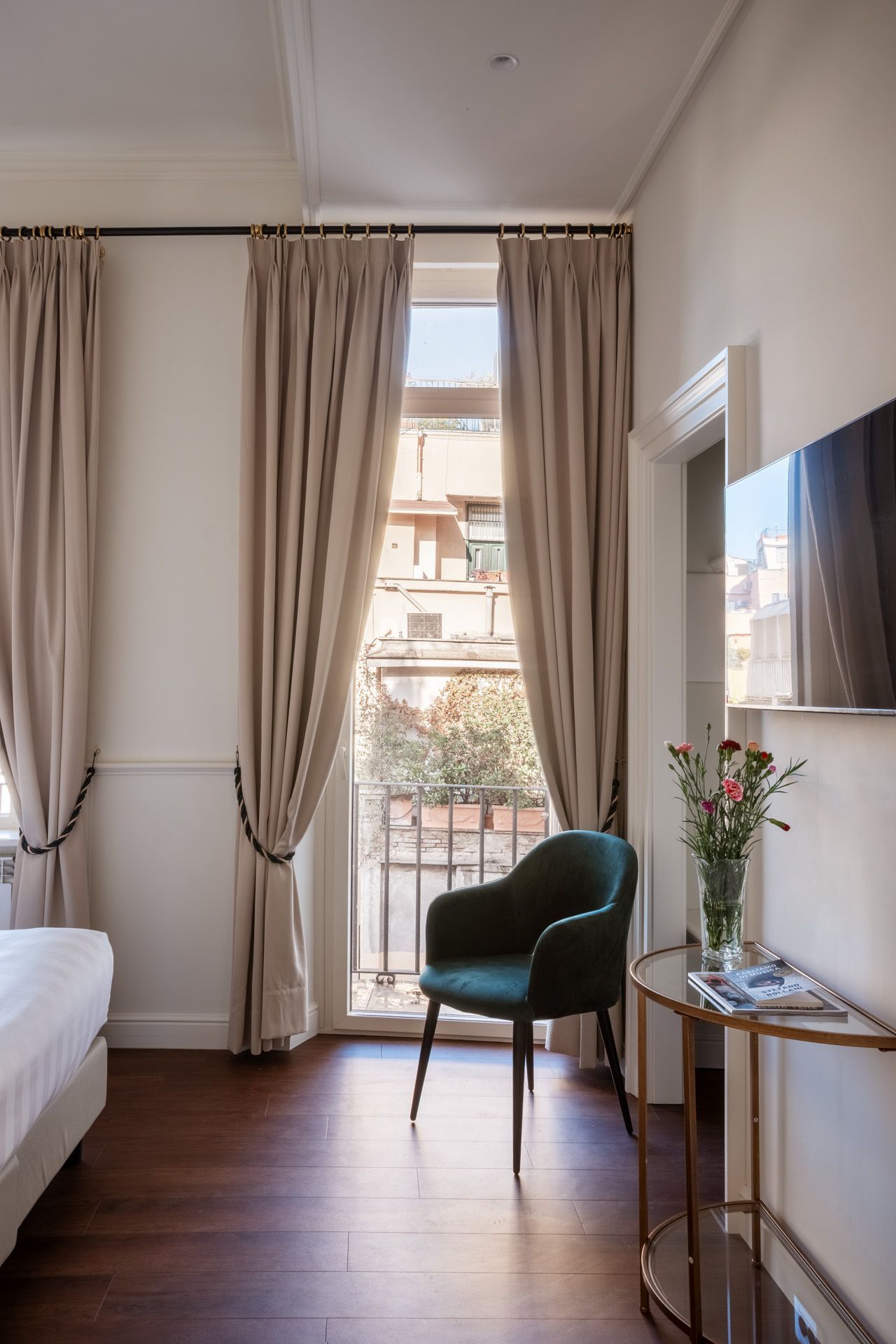 Room with balcony, green chair, tan curtains, small table with flowers, TV on wall.