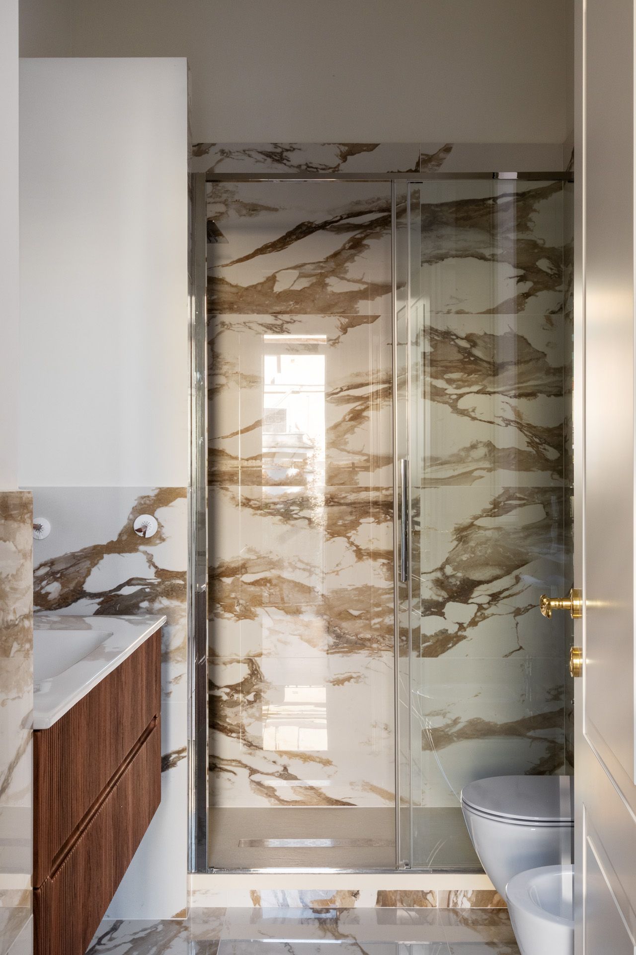 Modern bathroom with marble walls and glass shower. Features wooden vanity, toilet.