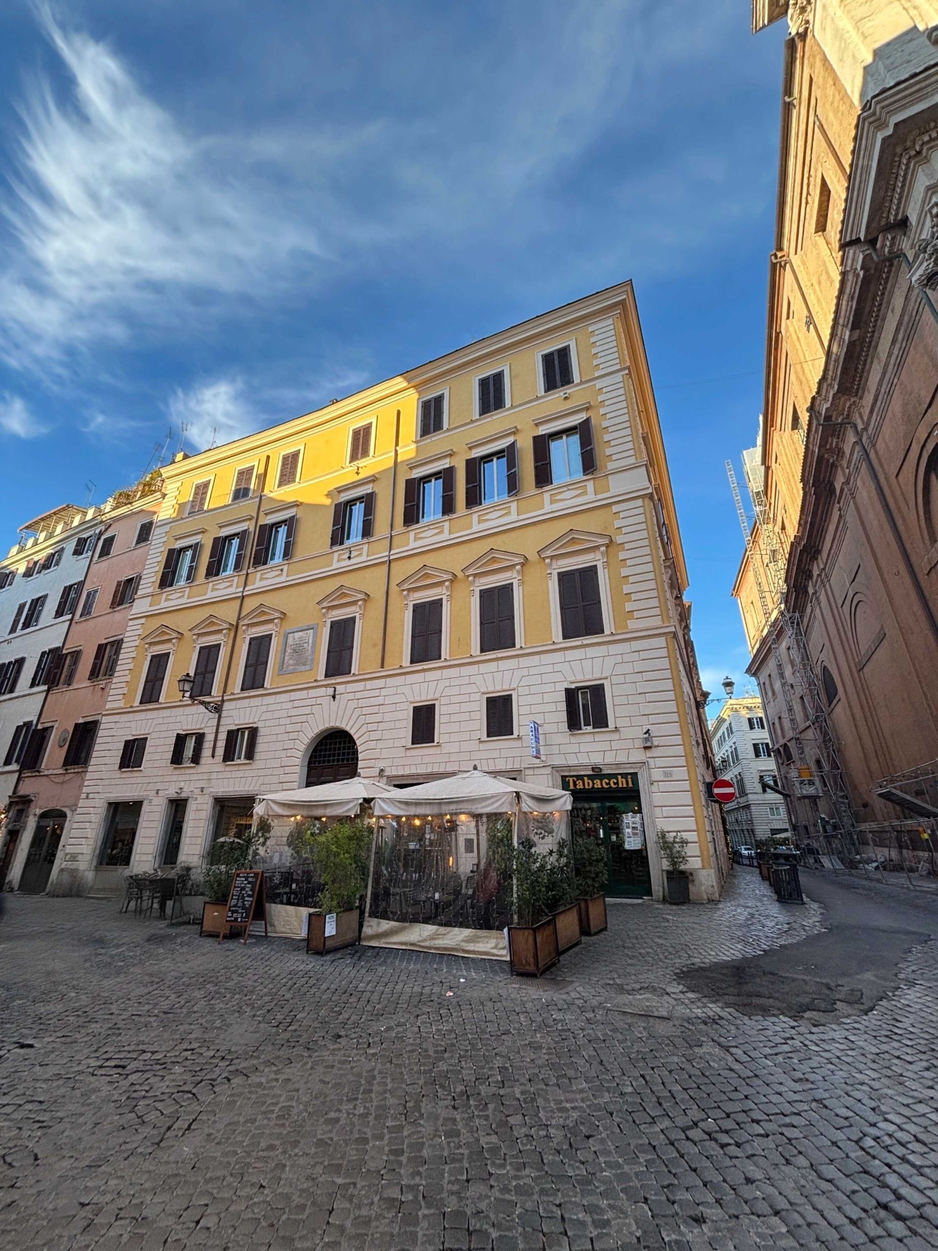 Buildings with a yellow facade on a cobblestone street under a blue sky, possibly in Italy.