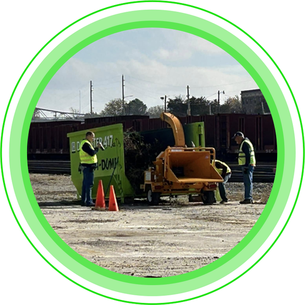 Men in safety vests operate a wood chipper near a green dumpster and train tracks.
