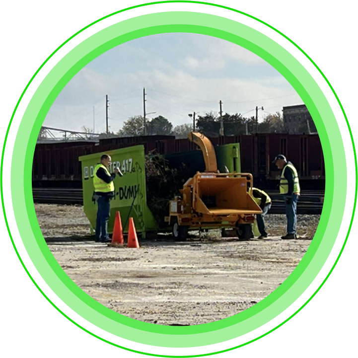 Men in safety vests operate a wood chipper near a green dumpster rented from dumpster417
