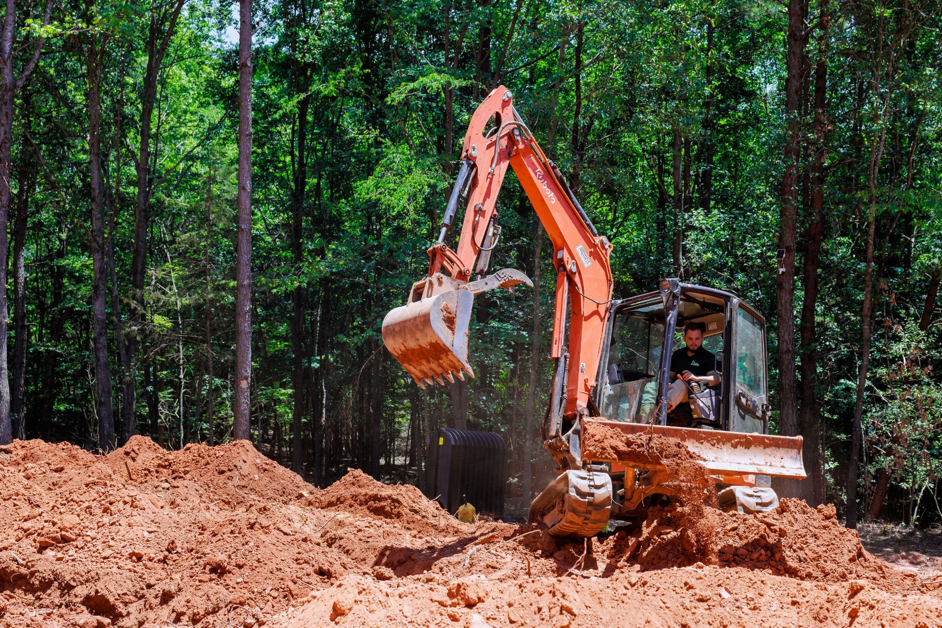 Orange excavator digging a large dirt pile in a wooded forest clearing