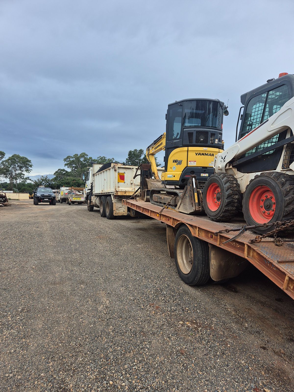 Flatbed Trailer Carrying Construction Machinery in a Gravel Lot Under Cloudy Skies — TP Concreting In Warilla, NSW