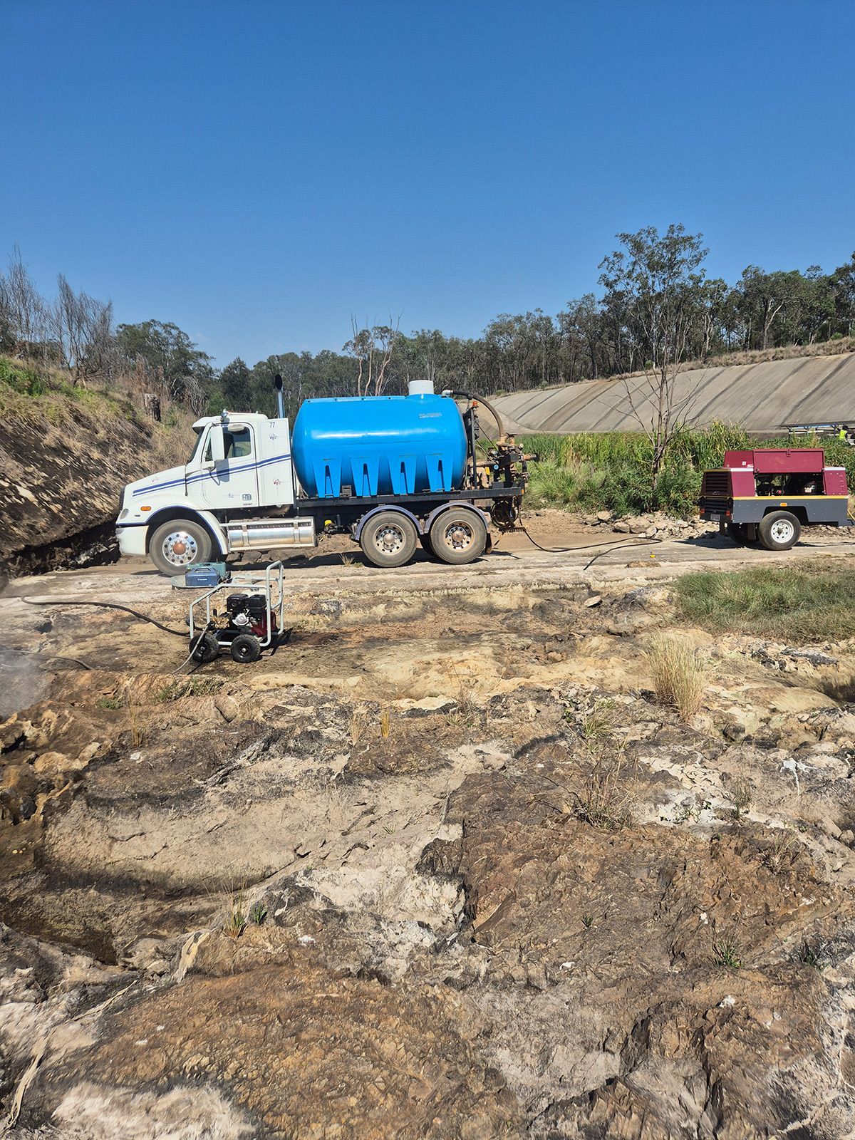 White Water Truck With Blue Tank and Red Trailer on Rocky Dirt Site Under Clear Sky — TP Concreting In Warilla, NSW
