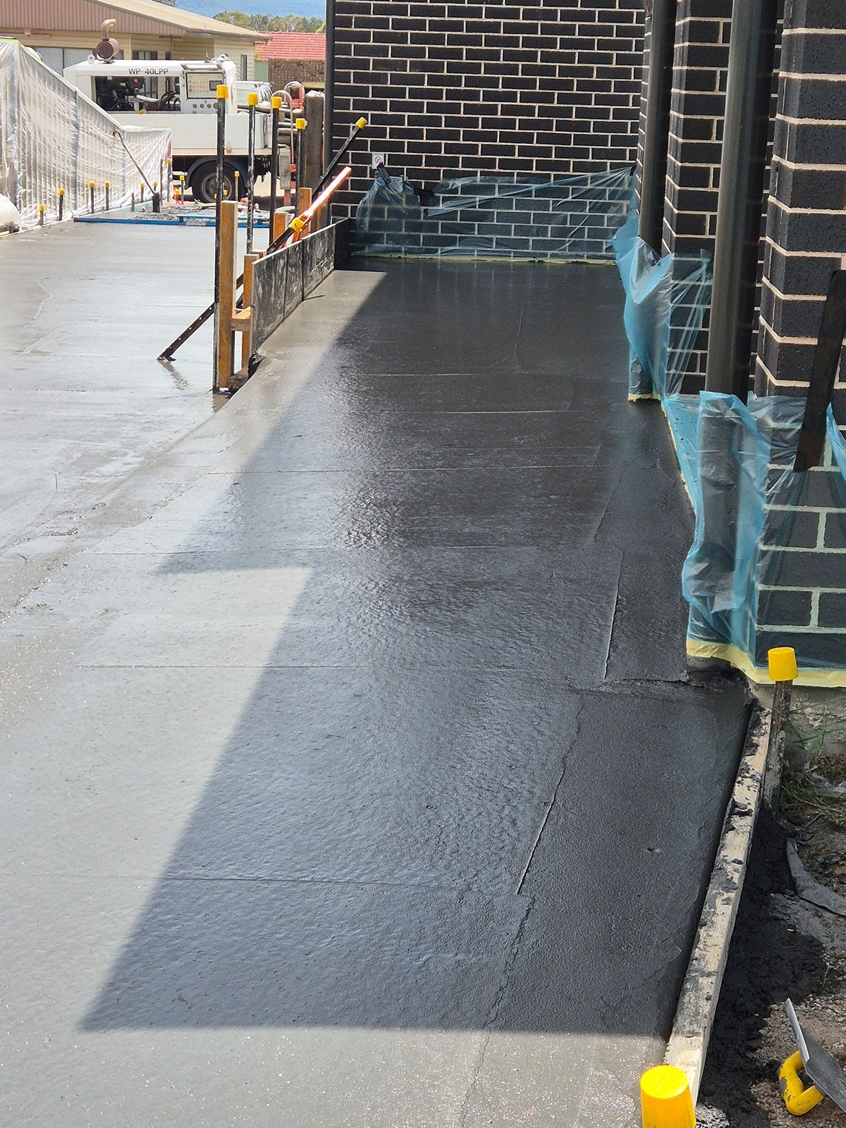 Freshly Coated Black Walkway Beside a Brick Building, With Blue Masking Tape and Safety Cones — TP Concreting In Warilla, NSW