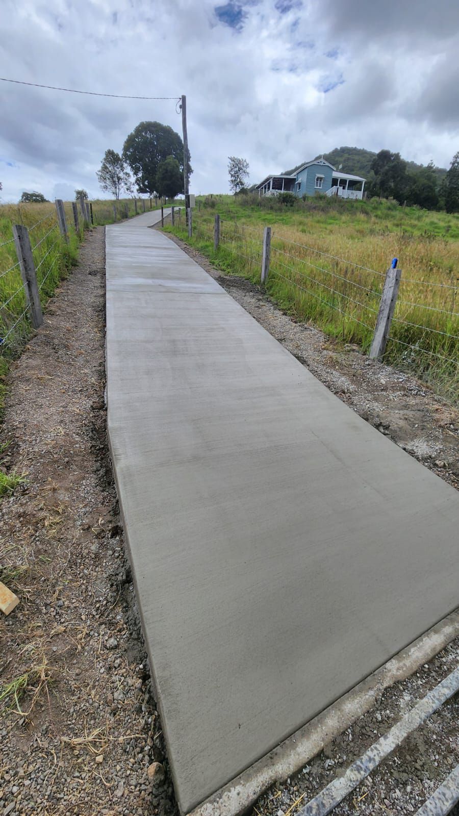 Fresh Concrete Walkway Along a Grassy Rural Roadside, Leading Toward a House and Trees — TP Concreting In Warilla, NSW