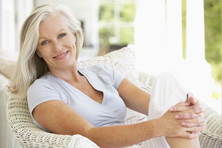 A woman is sitting in a wicker chair with her legs crossed and smiling.