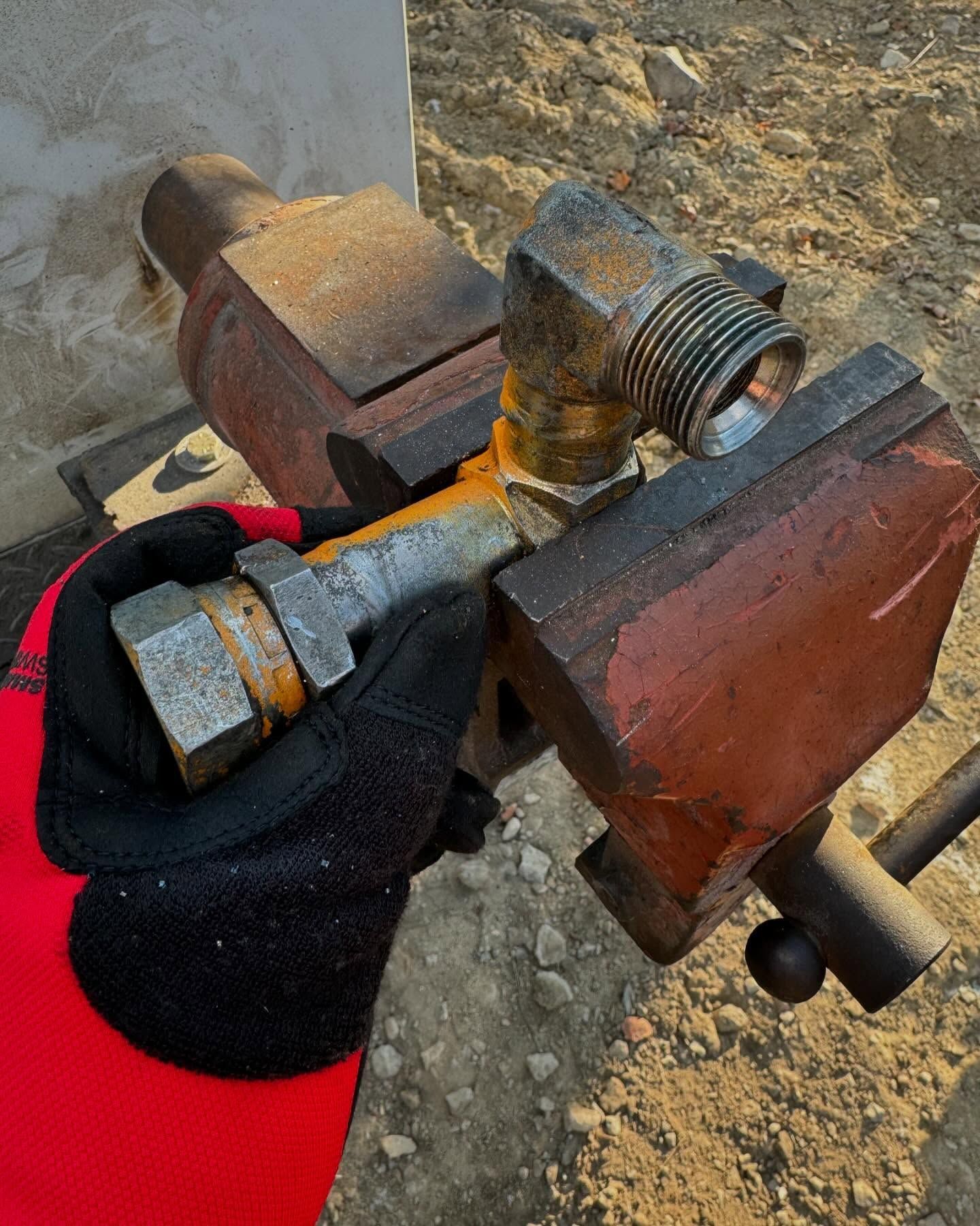 Gloved hand holding a rusted metal pipe fitting clamped in a bench vise outdoors.