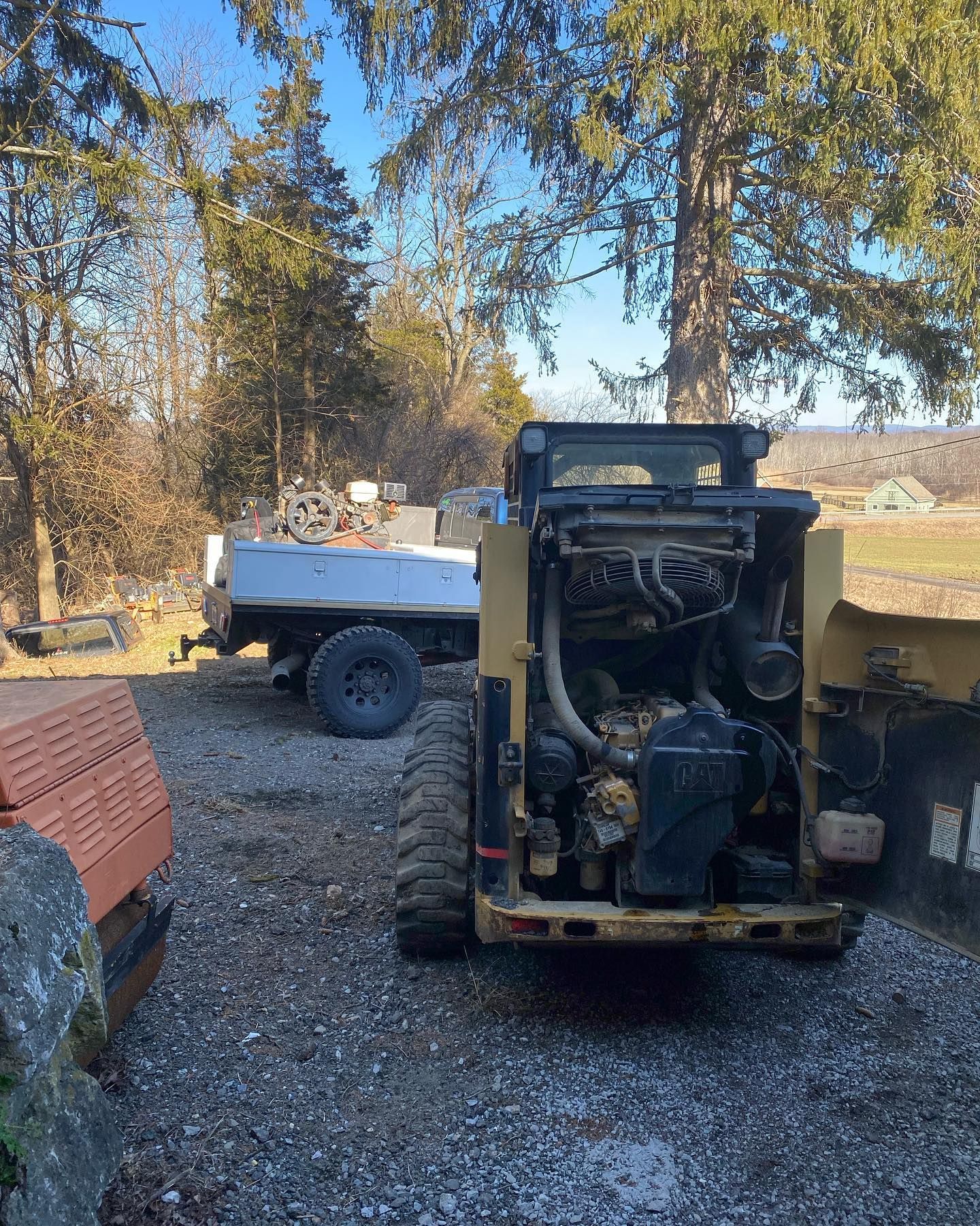 Old yellow skid-steer loader parked on gravel beside a white utility truck in a wooded yard.