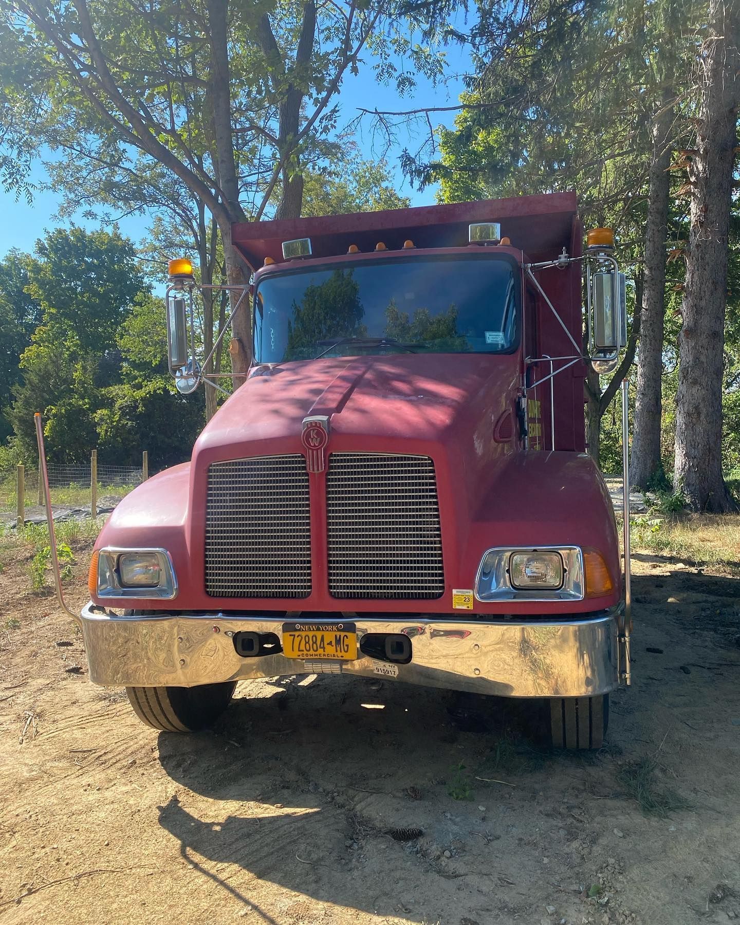 Red semi-truck parked outdoors on a dirt road among trees, facing the camera.